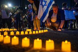 مردی کنار ردیفی از شمع ها زانو زده است. او پشت پرچم آبی و سفید اسرائیل است.